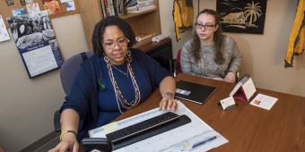 two woman sit at a desk. one of the woman is pointing at a monitor screen while the other listens on