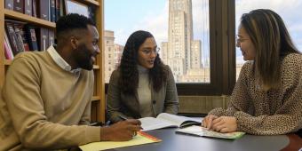 three people sit at a table talking. the cathedral of learning can be seen through the window behind them.